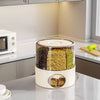 A cylindrical automatic grain dispenser with a cream-colored base and clear upper chamber, filled with black beans, yellow lentils, and green beans, set on a kitchen countertop.